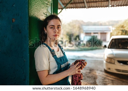 Gender equality. A portrait of a young brunette in uniform leaning against the gate and wiping her hands with a rag. In the background, a street and a white car