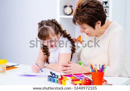 A girl with down syndrome draws at home next to the teacher. 