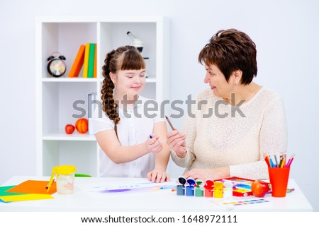 A girl with down syndrome draws at home next to the teacher. The teacher holds out a pencil to the girl