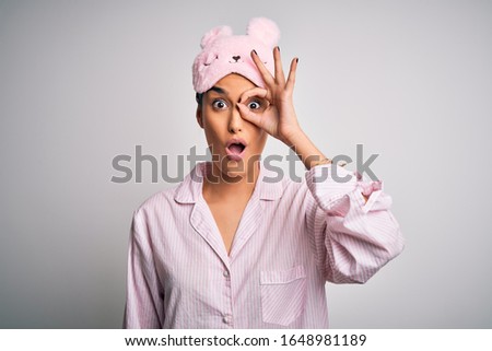Young beautiful brunette woman wearing pajama and sleep mask over white background doing ok gesture shocked with surprised face, eye looking through fingers. Unbelieving expression.