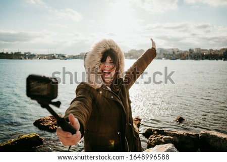 
Young tourist visiting a coastal town on the island of Malta. Taking pictures of herself with a small camera during a windy sunset wearing a coat. Lifesytle