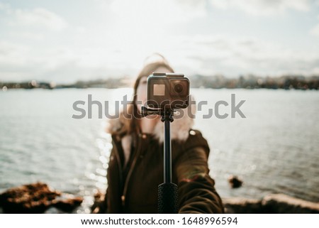 
Young tourist visiting a coastal town on the island of Malta. Taking pictures of herself with a small camera during a windy sunset wearing a coat. Lifesytle