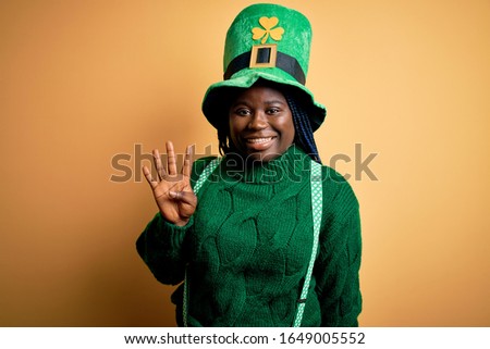 Plus size african american woman with braids wearing green hat with clover on st patricks day showing and pointing up with fingers number four while smiling confident and happy.