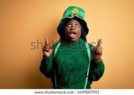 Plus size african american woman with braids wearing green hat with clover on st patricks day amazed and surprised looking up and pointing with fingers and raised arms.