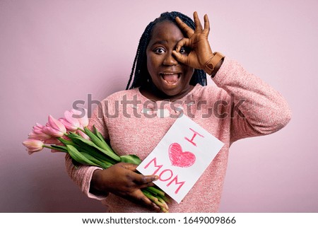 Plus size african american woman holding love mom message and tulips on mothers day with happy face smiling doing ok sign with hand on eye looking through fingers