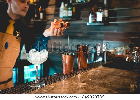 Young fancy sharp looking man bartender with yellow necktie and  apron making in process preparing alcoholic cocktail drink in restaurant bar on the counter table with bottles on the blur background