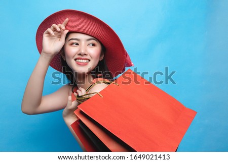 Portrait of young beautiful asian girl wearing dress and sun hat holding shopping bags and smile isolated over blue background. Online shopping and black friday concept.
