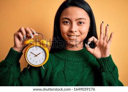 Young beautiful chinese woman holding vintage alarm clock over isolated yellow background doing ok sign with fingers, excellent symbol