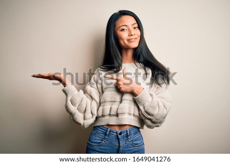 Young beautiful chinese woman wearing casual sweater over isolated white background amazed and smiling to the camera while presenting with hand and pointing with finger.