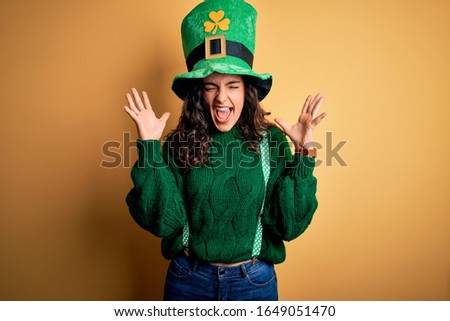 Beautiful curly hair woman wearing green hat with clover celebrating saint patricks day celebrating mad and crazy for success with arms raised and closed eyes screaming excited. Winner concept