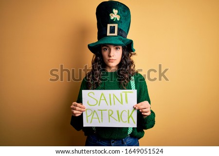 Beautiful curly hair woman wearing green hat holding banner with saint patricks message with a confident expression on smart face thinking serious