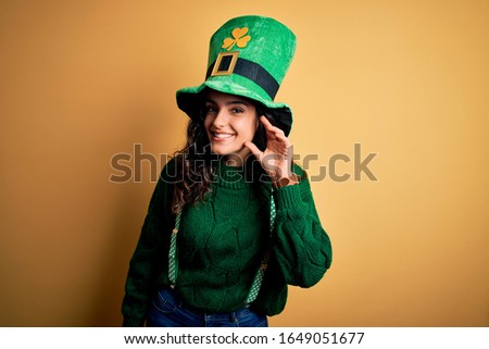 Beautiful curly hair woman wearing green hat with clover celebrating saint patricks day smiling with hand over ear listening an hearing to rumor or gossip. Deafness concept.