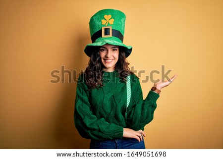 Beautiful curly hair woman wearing green hat with clover celebrating saint patricks day smiling cheerful presenting and pointing with palm of hand looking at the camera.