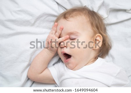 One year old baby girl boy wants to sleep, rubs his eyes with his hand and yawns on white sheets from above. Sleep training mode Royalty-Free Stock Photo #1649094604