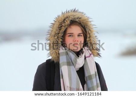 Beautiful young girl girl stands outdoors in winter clothes a jacket in the hood and a scarf on a background of a winter landscape with snow.