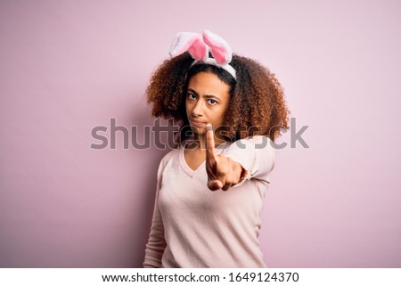 Young african american woman with afro hair wearing bunny ears over pink background Pointing with finger up and angry expression, showing no gesture