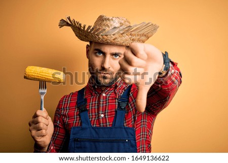Young rural farmer man holding countryside corn over isolated yellow background with angry face, negative sign showing dislike with thumbs down, rejection concept