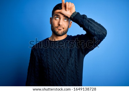 Young handsome man wearing casual sweater standing over isolated blue background making fun of people with fingers on forehead doing loser gesture mocking and insulting.