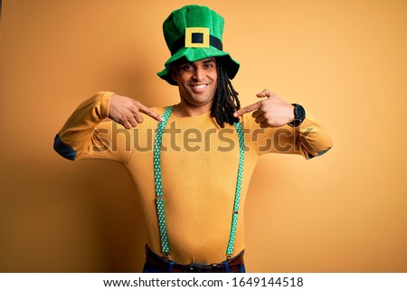 Young african american man wearing green hat celebrating saint patricks day looking confident with smile on face, pointing oneself with fingers proud and happy.
