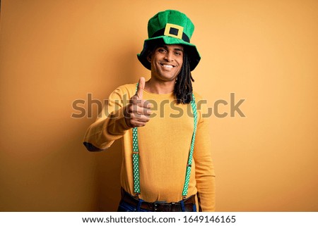 Young african american man wearing green hat celebrating saint patricks day doing happy thumbs up gesture with hand. Approving expression looking at the camera showing success.