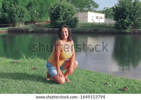 charming 50 year old woman with straight black hair, shoulder length, wearing dental braces, crouched on the grass beside a lake, happy