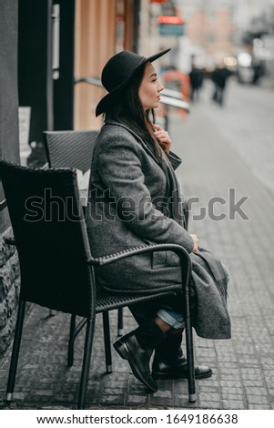 portrait of young beautiful romantic brunette woman in black hat, gray coat and scarf, jeans, sitting outdoors cafe happy in city on dark background