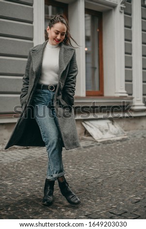 portrait of young beautiful brunette girl with high tail and red lipstick, gray coat, jeans, white sweater walks happy in the city on a background of gray classical architecture