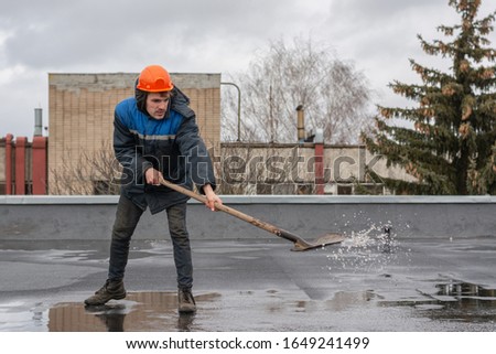 Worker builder on the roof of the factory removes water from the puddle with a shovel. on the head a construction helmet. work at height. Royalty-Free Stock Photo #1649241499