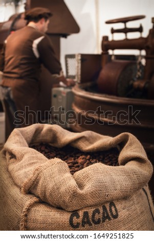 Jute bag full of cocoa beans in a chocolate maker workshop, with a male chocolatier working on conching and melanger equipment on the background Royalty-Free Stock Photo #1649251825