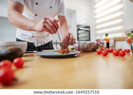 Cropped picture of dedicated caucasian chef standing in kitchen and putting cooked salmon on plate. On kitchen counter are different sorts of vegetables.