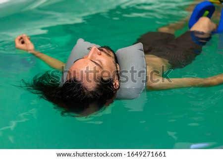 Young man with long hair and beard lying shirtless in swimming costume with eyes closed. relaxing in swimming pool with neck float for aqua therapy Royalty-Free Stock Photo #1649271661