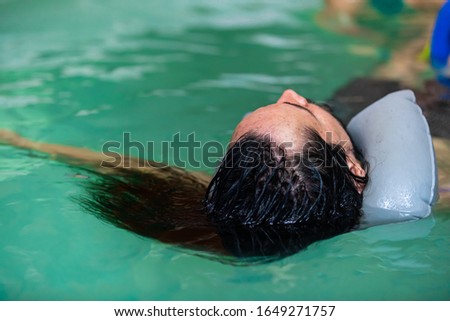 Close-up of young man with long hair floating on water in swimming pool feeling relaxed in spa centre during aqua therapy Royalty-Free Stock Photo #1649271757