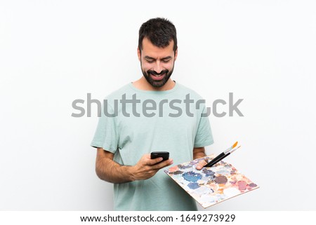 Young artist man holding a palette over isolated background sending a message with the mobile