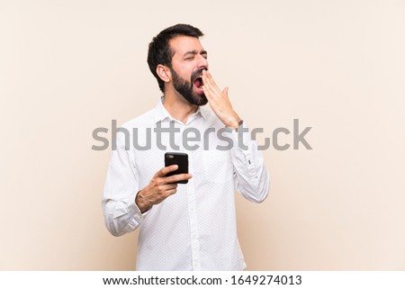 Young man with beard holding a mobile yawning and covering wide open mouth with hand