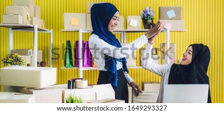 Muslim two asian woman freelancer sme business online shopping working on laptop computer with cardboard box on table at home - Business online shipping and delivery concept