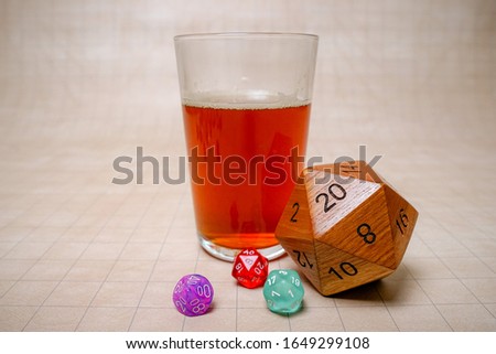 Beer in a pint glass near some big, wooden and colorful dice showing a twenty on a grid board game mat.