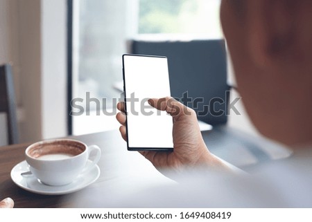 Mockup image of man hand holding and watching blank screen mobile smart phone, reading news via application with cup of coffee on wooden table in modern coffee shop, over shoulder view