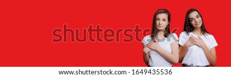 Two charming caucasian girls with black hair and beautiful smile are touching their hearts while advertising something on red freespaced background