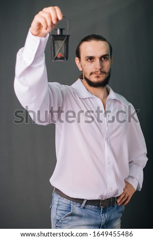 A guy with a beard in a white vintage shirt holds a black lantern. Romantic portrait on a gray background with a night lamp. Studio photo