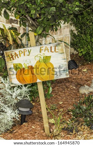 A sign reading Happy Fall standing in someones flower bed garden.