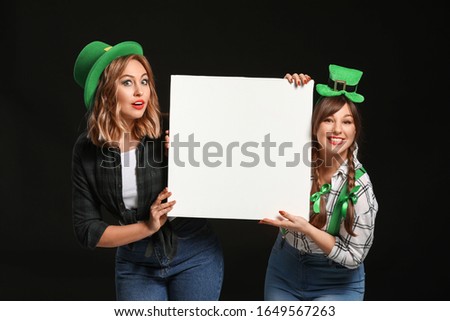 Funny young women with blank poster on dark background. St. Patrick's Day celebration