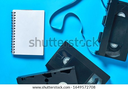 Scattered many black old plastic vhs video cassettes near blank notepad lies on blue desk. Concept of 90s. Copy space. Top view