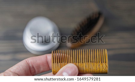 Wooden Beard Comb In A Male Hand On A Blurred Background Of A Wooden Table. Hairdressing Supplies. High Quality Photo