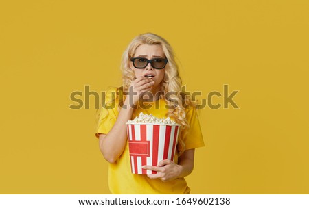 Surprised young blond woman in 3d glasses and yellow shirt eating popcorn, looks shocking movie at the cinema. Isolated on yellow background