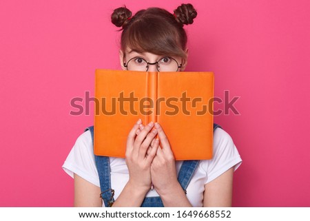 Horizontal shot of winsome young student with funny hair buns, girl hiding behind orange book, diary or planner, female wearing glasses, white t shirt, red bandana over neck and denim overalls.