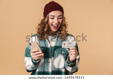 Photo of nice cheerful woman in knit hat holding credit card and cellphone isolated over beige background
