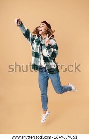 Photo of pretty joyful woman in knit hat taking selfie and gesturing peace sign while jumping isolated over beige background