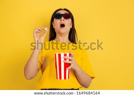 Watching cinema in 3D-eyewear with popcorn. Caucasian woman on yellow studio background. Beautiful brunette model in casual style. Concept of human emotions, facial expression, sales, ad, copyspace.