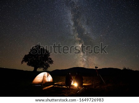 Summer night camping, tourism and photography. Back view of two men sitting on big logs between tourist tent and tripod camera at burning campfire under dark starry sky with Milky Way constellation.