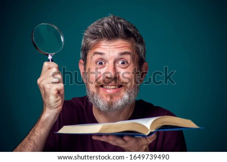 A portrait of bearded man in red t-shirt holding opened book and magnifying glass. People, education and searching concept